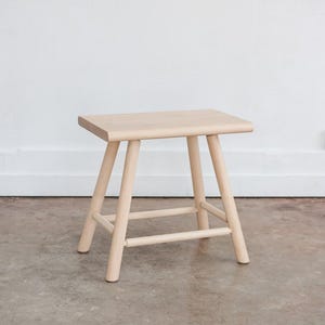 May include: A simple wooden stool with a rectangular seat and four legs. The stool is made of light wood and has a natural finish.