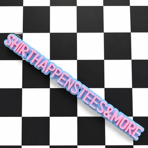 May include: A pink and blue sign that reads "SHIRTHAPPENSTEES&MORE" is displayed diagonally across a black and white checkered background. The sign has a light blue outline and pink lettering.