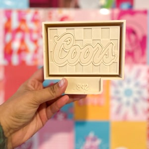 May include: A cream-colored Coors logo mold with a checkered background. The word "Coors" is in a cursive font. The number "892" is at the bottom of the mold. The mold is held in a hand against a colorful background.