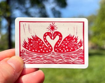 May include: A rectangular sticker featuring a red illustration of two swans forming a heart shape, with a starburst above. The swans are decorated with star patterns and sit on wavy lines. The sticker has a white background with a red border.