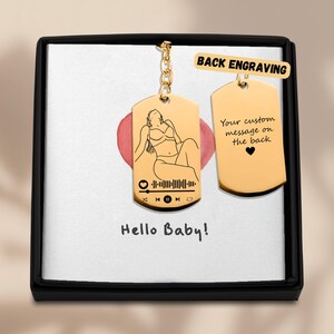 May include: Gold keychain with a personalized engraved tag featuring a line drawing of a woman and a music player icon with a barcode. The back of the tag has the text "Your custom message on the back". The keychain is in a black box with a white background.