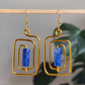 May include: A pair of gold-toned wire earrings featuring a rectangular spiral frame design. Each earring suspends a single, cylindrical, marbled blue bead in the center, attached to a gold-toned hook for hanging, creating a geometric aesthetic.