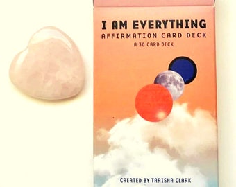 May include: A pink heart-shaped rose quartz crystal next to a box of affirmation cards. The box is pink, orange, and blue with the text "I AM EVERYTHING AFFIRMATION CARD DECK A 30 CARD DECK CREATED BY TARISHA CLARK".