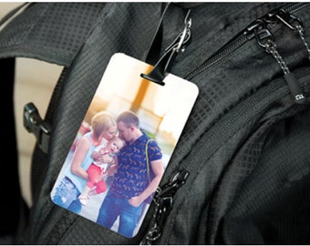 May include: A rectangular luggage tag featuring a printed colour photograph of a family of three, including two adults and a small child, attached to a black textured backpack with a black plastic strap and a metal clip mechanism.