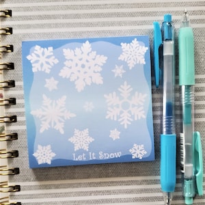 May include: A square blue sticky note with white snowflakes and the text "Let It Snow." Two pens, one blue and one mint green, are next to the sticky note. The background is a notebook with a striped pattern.