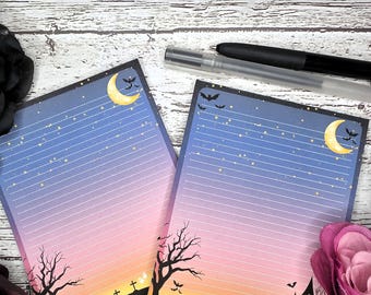 May include: Two Halloween-themed notepads with a gradient sky design, featuring a crescent moon, bats, and a spooky silhouette of a haunted house, trees, and tombstones. Lined pages with a black pen on a wooden surface.