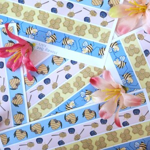 Peut inclure: Un ensemble de quatre rubans washi sur le thème de l'abeille. Les rubans sont bleus, jaunes, roses et blancs avec un motif répétitif de nids d'abeilles, d'abeilles et de louches à miel. Le texte sur les rubans indique "Honey Bee Washi Tape", "Kids Stationery Service" et "Honey Bee's Stickers".
