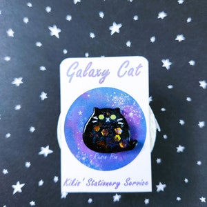 May include: A black resin pin of a cat with a galaxy design. The cat is sitting on a white background with a purple and blue galaxy design. The pin is labeled "Galaxy Cat" and "Kiki's Stationery Service".