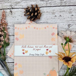 May include: A 4x5 inch memo pad with a beige background and a grid pattern. The pad has an autumnal theme with orange, yellow, and red leaves and pumpkins. The text on the pad reads "Hello Autumn: 4x5 inch Memo pad 30 pages Dreamy Cottage Crafts".