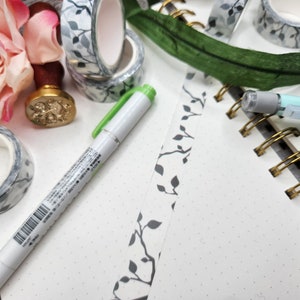 Op de afbeelding: Een wit notitieboek met een gouden spiraalbinding en een rand van grijs-witte gepatenteerde washi-tape. Een groene pen en een rol grijs-witte washi-tape zijn ook zichtbaar.