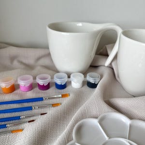 Könnte beinhalten: Ein Satz mit Farbtöpfen mit orangefarbener, rosa, lila, blauer und weißer Farbe, zusammen mit Pinseln und einer weißen Keramik-Farbpalette. Im Hintergrund befinden sich zwei weiße Keramikbecher mit Henkeln.