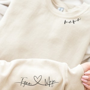 Könnte beinhalten: Ein beiger Pullover mit dem Text "mama" oben und "Fynn & Maja" mit einem Herzsymbol auf dem Ärmel.