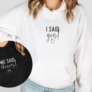 May include: Two white hoodies with black text. One hoodie says "I said yes!" with a ring symbol. The other hoodie says "We said cheers!" with a champagne glass symbol.