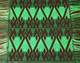 Pode incluir: Um corredor de mesa de crochê verde e marrom com franjas nas pontas. O corredor apresenta um padrão geométrico de diamantes e triângulos.