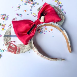 May include: Red satin bow with a fork and cork tucked into the bow. The bow is attached to a headband with a white and brown patterned fabric. The headband is decorated with a map-like design.
