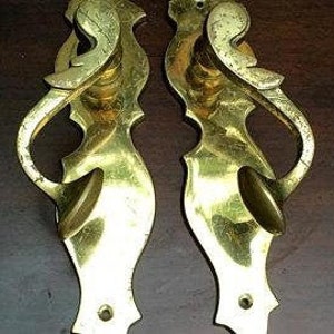 K&ouml;nnte beinhalten: Ein Paar antike Messing-T&uuml;rgriffe mit verzierten, dekorativen Designs. Die Griffe haben eine einzigartige, geschwungene Form und eine glatte, polierte Oberfl&auml;che.