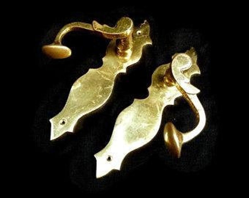 K&ouml;nnte beinhalten: Zwei verzierte Messing-T&uuml;rgriffe mit einem dekorativen Design. Die Griffe haben eine gebogene Form und einen kleinen Knopf am Ende.