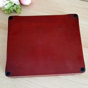 May include: A rectangular, reddish-brown wooden box with rounded edges and four small black feet. The box has a smooth, polished surface. A few flowers are visible in the upper left corner.