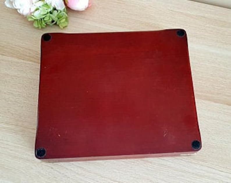May include: A rectangular, reddish-brown wooden box with rounded edges and four small black feet. The box has a smooth, polished surface. A few flowers are visible in the upper left corner.