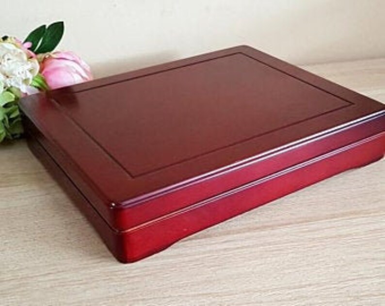May include: A rectangular, reddish-brown wooden box with a glossy finish. The box has a slightly raised, darker border around the lid. A few pink and white flowers with green leaves are visible in the background.