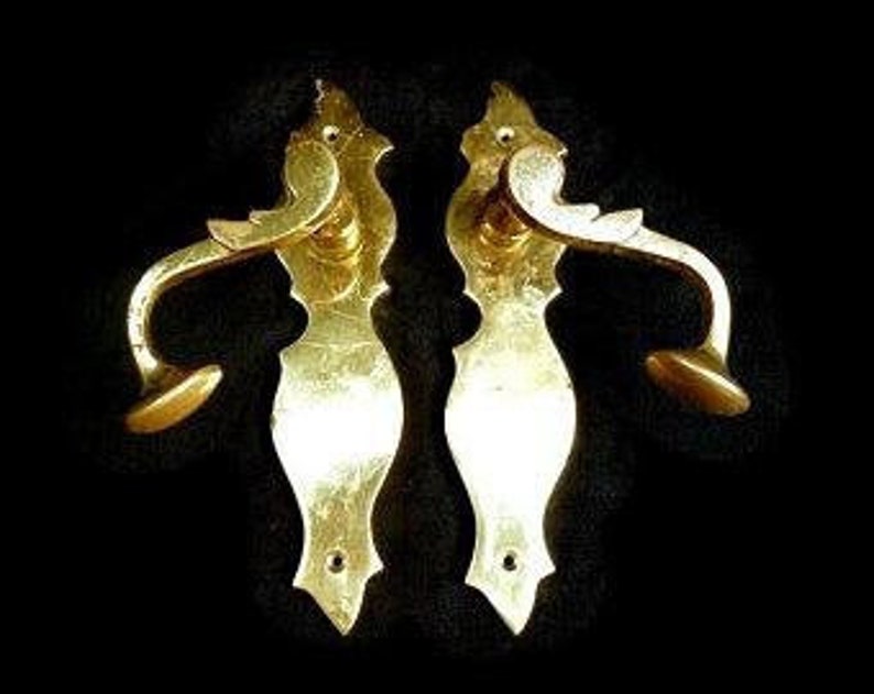 K&ouml;nnte beinhalten: Ein Paar verzierte goldene T&uuml;rklinken mit einem dekorativen, flie&szlig;enden Design. Die Griffe haben ein Hebeldesign und einen runden Knopf.