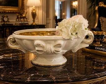Op de afbeelding: Een witte keramische centerpiece kom met gouden randen en strikdetails, gevuld met witte bloemen. De kom heeft een gouden interieur en staat op een donker marmeren oppervlak. Decoratief en elegant.