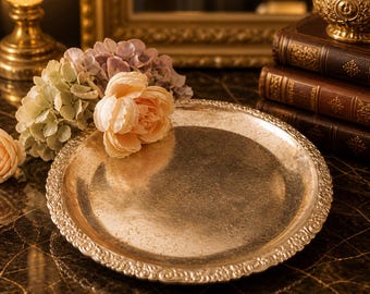 May include: A round, antique-style silver tray with an ornate, scrolled edge. The tray is displayed on a dark, patterned surface and is adorned with peach and lavender flowers. A gold-colored lamp and books are in the background.