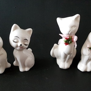 Puede incluir: Cuatro figuras de gato de cerámica blanca con los ojos cerrados y detalles pintados. Dos gatos tienen lazos rosas con vegetación, mientras que los otros dos tienen pestañas pintadas y labios rojos. Las figuras están sobre una superficie negra.