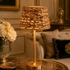 May include: A table lamp with a slender gold-toned metal stand and a circular base. The lamp features a conical lampshade crafted from woven natural fibres, which emits a warm, diffused light through the gaps in the braided material.