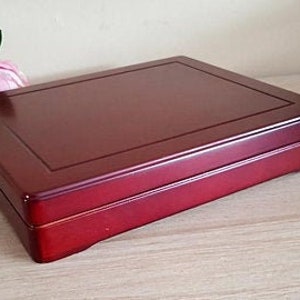 May include: A rectangular, lacquered wooden box in a deep red hue. The box features a slightly raised, rectangular border on the lid and a clean, minimalist design. The box is likely used for storage or display of small items.