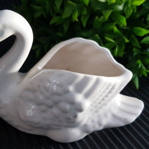 Puede incluir: Un macetero de cerámica blanca en forma de cisne con un diseño detallado. El cisne tiene la boca abierta, formando un macetero para plantas o flores pequeñas.