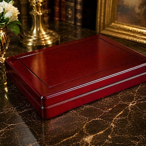 May include: A rectangular, wooden box with a rich, dark red finish. The box has a flat top with a slightly raised border and a polished surface. The box is likely for storing jewelry or other small items. The box is sitting on a marble surface.