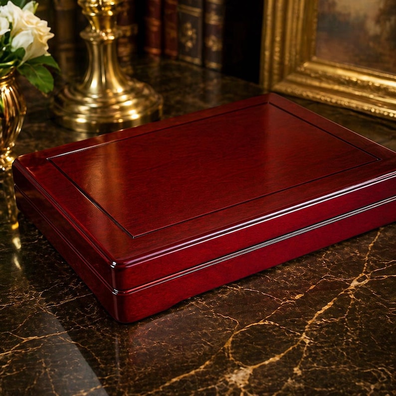 May include: A rectangular, wooden box with a rich, dark red finish. The box has a flat top with a slightly raised border and a polished surface. The box is likely for storing jewelry or other small items. The box is sitting on a marble surface.