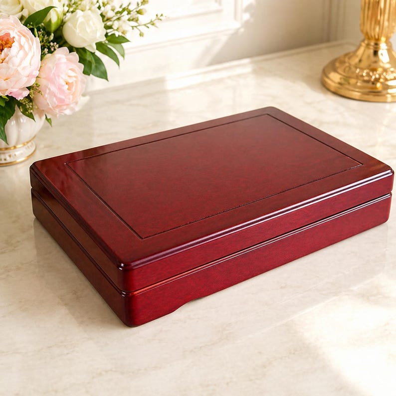May include: A rectangular, wooden box with a rich, dark red finish. The box has a glossy surface and a slightly raised, framed top. The design suggests a storage or display case, possibly for jewelry or keepsakes.