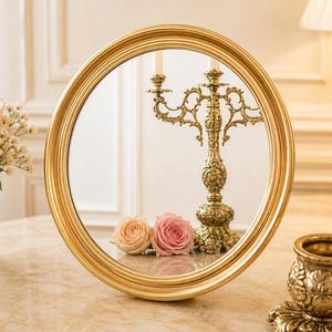 May include: A round, gold-framed mirror reflects a gold candelabra with lit candles and two roses. The mirror sits on a marble-like surface, with a small gold decorative piece in the foreground. The overall theme is vintage decor.
