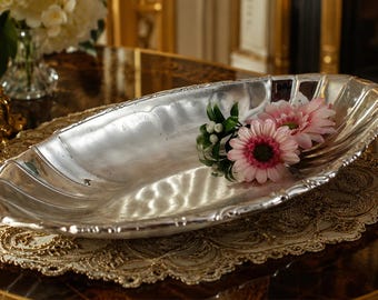 Könnte beinhalten: Eine silberfarbene, längliche Servierschale mit gewelltem Rand, die auf einer dekorativen Deckchen liegt. Die Schale enthält rosa und weiße Blumen. Der Hintergrund ist ein Tisch mit goldfarbenem Rand.