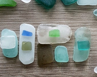 May include: Assortment of sea glass pieces in various shapes and colors, including blue, green, and white. The sea glass is arranged on a wooden surface.