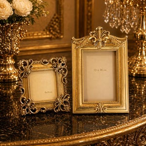May include: Two ornate gold picture frames. The smaller frame is 9 x 9 cm, and the larger frame is 13 x 18 cm. Both frames have intricate floral and scrollwork designs. The frames are displayed on a dark, glossy surface.