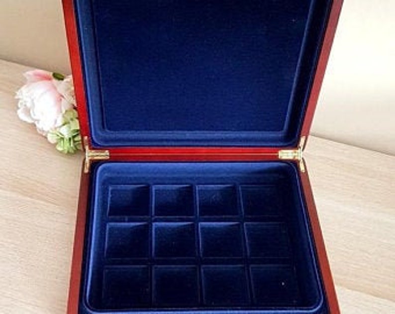 May include: Open wooden jewelry box with a red exterior and a deep blue velvet interior. The box has a rectangular shape with a hinged lid and gold-colored hardware. The interior features a grid of square compartments.
