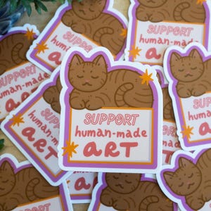 Puede incluir: Una colecci&oacute;n de pegatinas con un gato atigrado marr&oacute;n descansando sobre un letrero. El letrero dice "SUPPORT human-made ART" en texto rosa y blanco, con acentos de estrellas naranjas. Las pegatinas tienen un borde blanco con un contorno morado.