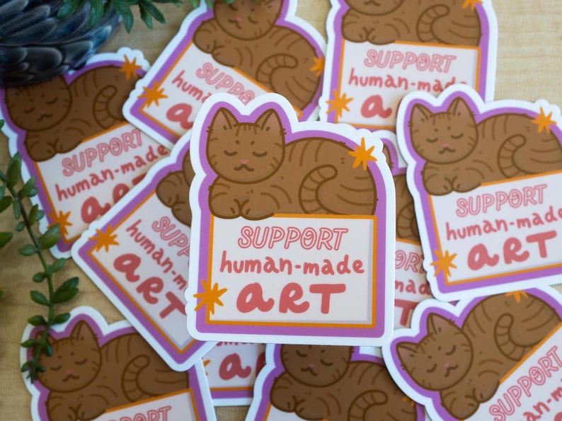 Puede incluir: Una colecci&oacute;n de pegatinas con un gato atigrado marr&oacute;n descansando sobre un letrero. El letrero dice "SUPPORT human-made ART" en texto rosa y blanco, con acentos de estrellas naranjas. Las pegatinas tienen un borde blanco con un contorno morado.