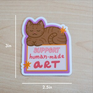 Puede incluir: Adhesivo con una ilustraci&oacute;n de gato atigrado marr&oacute;n, de 7,6 cm por 6,4 cm. El adhesivo tiene el texto "SUPPORT human-made ART" en rosa y blanco. El gato est&aacute; acostado, con un borde morado y naranja.