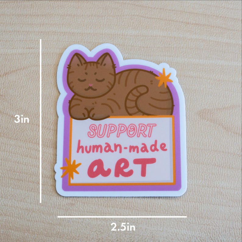 Puede incluir: Adhesivo con una ilustraci&oacute;n de gato atigrado marr&oacute;n, de 7,6 cm por 6,4 cm. El adhesivo tiene el texto "SUPPORT human-made ART" en rosa y blanco. El gato est&aacute; acostado, con un borde morado y naranja.