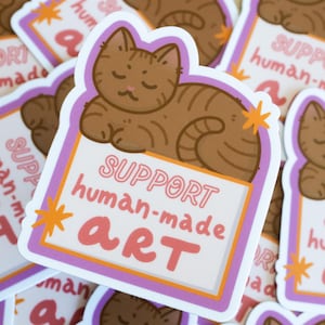 Puede incluir: Una pegatina con un gato atigrado marr&oacute;n descansando sobre un letrero. El letrero dice "SUPPORT human-made ART" en letras rosas y naranjas. La pegatina tiene un fondo blanco con un borde morado y naranja. Varias pegatinas similares son visibles.