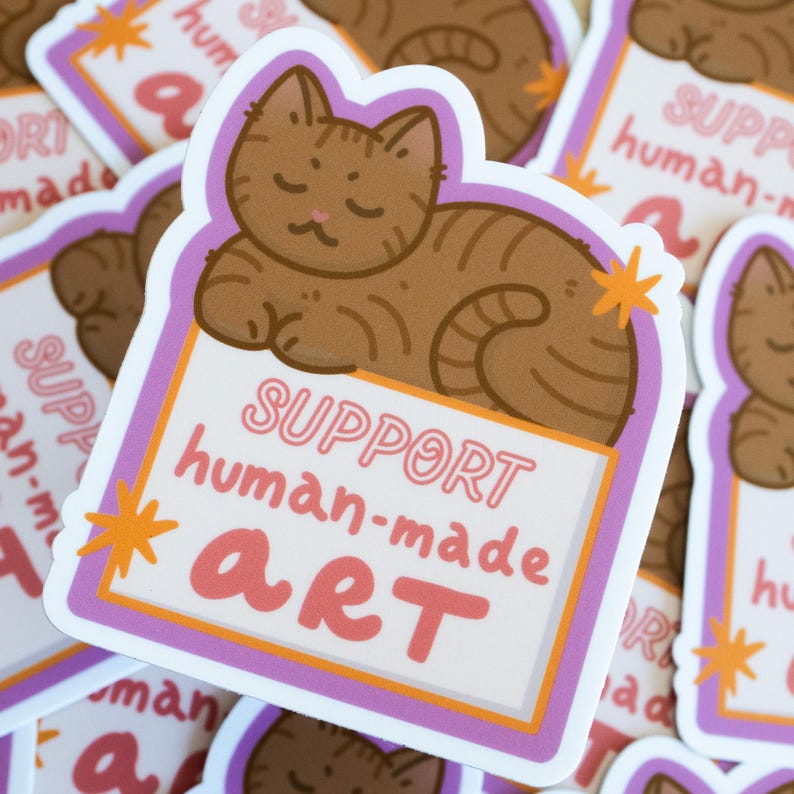 Puede incluir: Una pegatina con un gato atigrado marr&oacute;n descansando sobre un letrero. El letrero dice "SUPPORT human-made ART" en letras rosas y naranjas. La pegatina tiene un fondo blanco con un borde morado y naranja. Varias pegatinas similares son visibles.