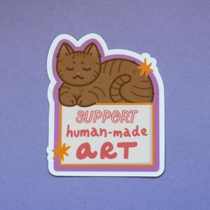 Puede incluir: Una pegatina con un gato atigrado marr&oacute;n tumbado sobre un letrero. El letrero dice "SUPPORT human-made ART" en rosa y naranja. La pegatina tiene un borde blanco y un fondo morado.