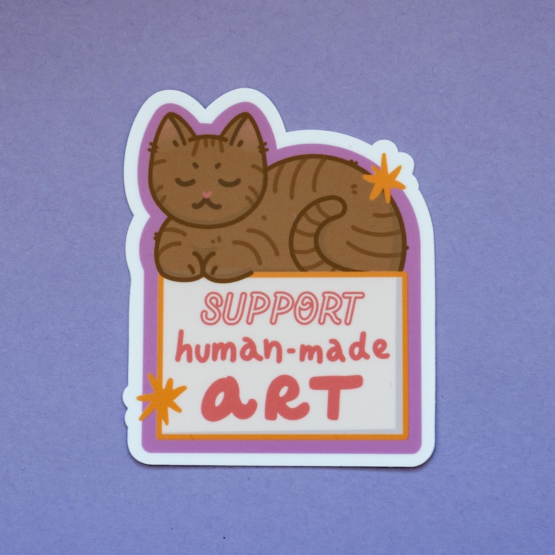 Puede incluir: Una pegatina con un gato atigrado marr&oacute;n tumbado sobre un letrero. El letrero dice "SUPPORT human-made ART" en rosa y naranja. La pegatina tiene un borde blanco y un fondo morado.