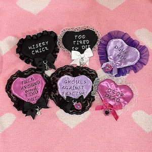 May include: Six heart-shaped brooches with various phrases, including "Misery Chick" and "Too Tired to Die." Each brooch has a different color, lace trim, and bow. The background is a pink sweater with white heart patterns.