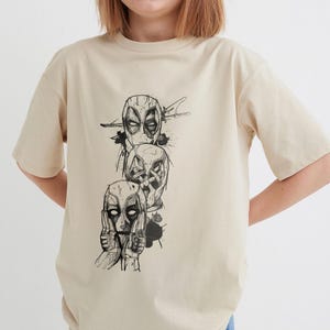May include: A beige t-shirt with a black and white graphic print of three Deadpool characters. The characters are stacked on top of each other, with the top character's eyes closed and the bottom character's hands covering their ears.