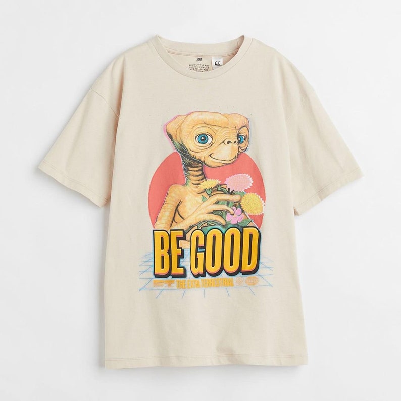 Pode incluir: Camiseta bege com uma estampa gr&aacute;fica de E.T. o extraterrestre segurando flores. O texto "BE GOOD" est&aacute; impresso abaixo da imagem.